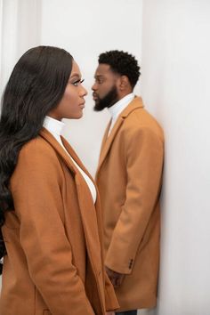 a man and woman standing next to each other in front of a white wall, looking at each other