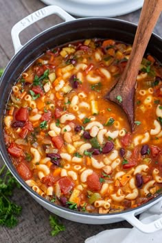 a pot filled with pasta and beans on top of a wooden table next to a spoon