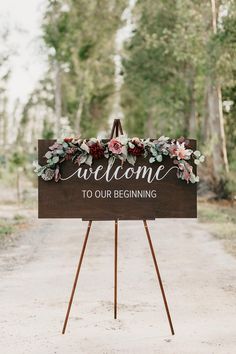 a welcome sign with flowers and greenery on it is standing in the middle of a dirt road