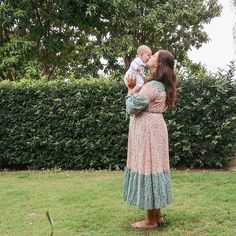 a woman holding a baby in her arms while standing on top of a lush green field