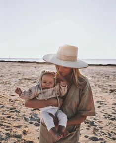 a woman holding a baby in her arms on the beach while wearing a cowboy hat