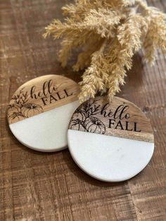 two wooden coasters sitting on top of a table next to a dried plant with the words hello fall written on them