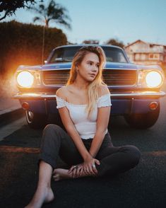 a woman sitting on the ground in front of a car with her legs crossed and head turned to the side