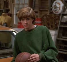 a young man holding a basketball in front of a parked car and looking at the camera