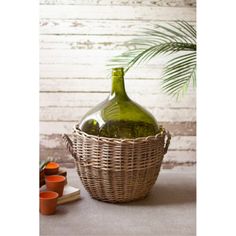 a green glass bottle in a wicker basket next to two cups and a plant