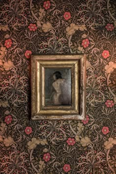 a painting hanging on the wall in front of a flowered wallpaper with a woman's reflection