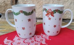 two coffee mugs decorated with holly and bows on red table cloth next to stone wall