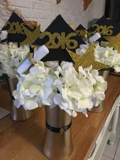 three graduation hats with flowers in them on a wooden table next to white and gold decorations