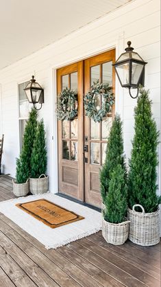 two potted trees sitting on the front porch next to a door with wreaths