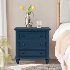 a blue dresser with two drawers and a vase on the top, next to a bed