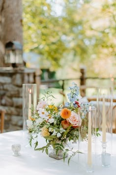 an arrangement of flowers and candles on a white table cloth with wood chairs in the background