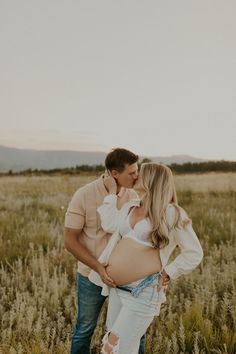 a pregnant woman kissing her husband while standing in the middle of a field with tall grass