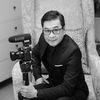 black and white photograph of man sitting on chair with camera attached to arm, smiling at camera