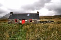 an old stone house with a red door in the middle of tall grass and mountains