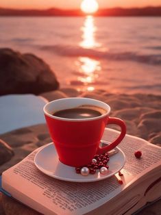 a cup of coffee sitting on top of an open book next to the ocean at sunset