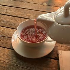 a teapot pouring red liquid into a white cup on a wooden table next to a napkin