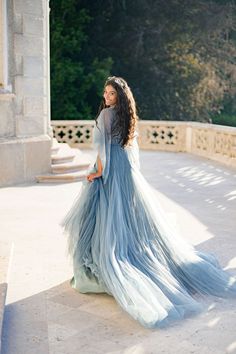 a woman in a long blue dress standing on a stone walkway with trees in the background