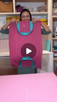 a woman holding up a pink and blue baby carrier over a table with shelves in the background