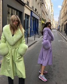 two women are walking down the street in coats and fur stoles, one is wearing a purple coat