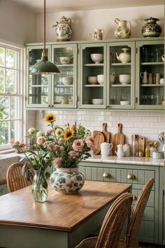 a kitchen with green cabinets and flowers in a vase on the counter top, surrounded by wicker chairs