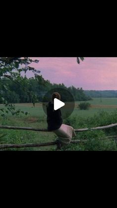 a woman sitting on top of a wooden fence next to a lush green field at sunset