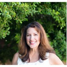 a woman with brown hair and white shirt smiling in front of green leaves on a tree