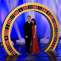 a man and woman standing in front of a large clock with numbers on it's sides