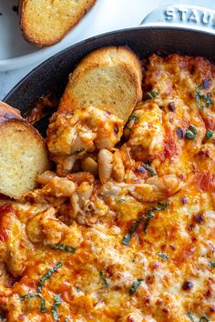 a skillet filled with cheese and bread on top of a white table next to garlic bread