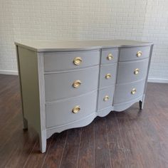 a grey dresser with gold handles on it's drawers and wood floors in front of a white brick wall