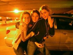 three young women standing next to each other in front of a parked car at night