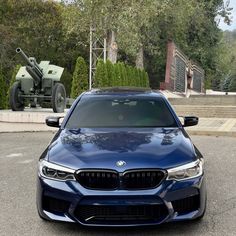 a blue bmw is parked in front of a large tank on the street next to some bushes and trees