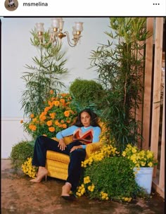 a woman sitting on a chair in front of some flowers and potted plants, reading a book