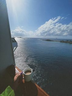 a cup of coffee sitting on top of a wooden table next to the ocean under a blue sky