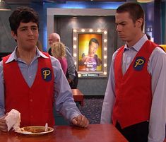 two men in red vests are standing at a table and one is eating food