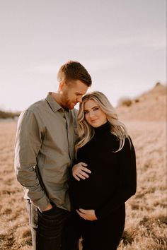 a pregnant couple standing in a field with their arms around each other and looking at the camera