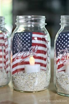 three mason jars filled with sand and a lit candle on top of the jar is decorated with an american flag