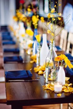 a long table with yellow flowers and vases filled with water, candles and napkins