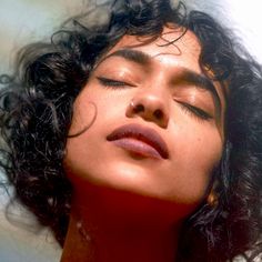 a woman with her eyes closed wearing a red shirt and black curly hair is looking up at the sky