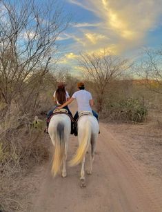 two people riding horses down a dirt road in the middle of trees and bushes on either side