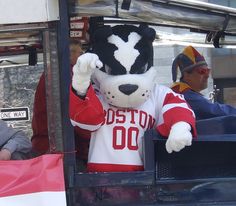 a mascot in the back of a vehicle waving to someone onlooker behind him