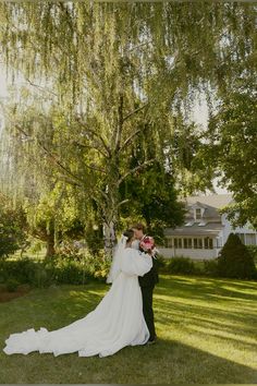 a bride and groom standing under a tree in front of a large white house on a sunny day