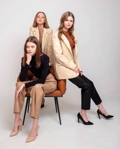 three women sitting on chairs in front of a white background, one is wearing a tan coat and the other has black pants