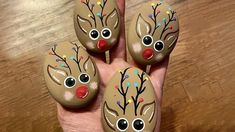 four painted rocks with reindeer faces on them sitting on a wooden table next to a person's hand