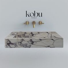 a bathroom sink with two gold faucets on the wall and a kobu logo above it