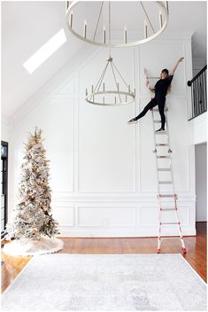 a woman climbing up the side of a ladder next to a christmas tree in a white room