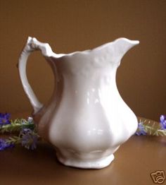 a white pitcher sitting on top of a table next to blue and purple flowers in front of a brown wall