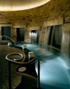 an indoor swimming pool surrounded by stone walls and water features, including a spiral staircase