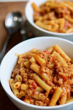 two white bowls filled with pasta and meat on top of a wooden table next to spoons