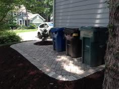 three trash cans sitting next to a tree in front of a house on a brick walkway