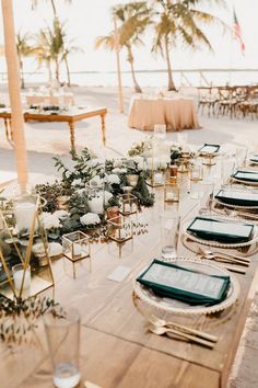 a table set up with place settings and greenery for an outdoor wedding reception on the beach